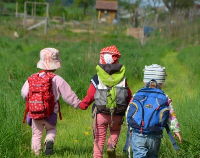 drei Kinder mit Rucksäcken von hinten. Sie gehen über eine Wiese in Richtung Gemüsegarten. Zwei der Kinder geben einander die Hand.
