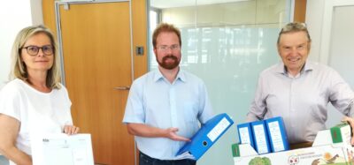 eine Frau und zwei Männer stehen in einem Büro, in der Hand mehrere Ordner bzw. eine ganze Schachtel mit Ordnern