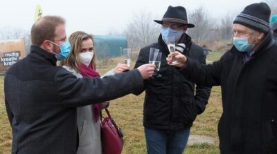 drei Männer und eine Frau stoßen mit Sektgläsen an. Sie tragen die obligatorischen Masken. Im Hintergrund eine Wiese, Bauzäune und kahle Sträucher im Nebel