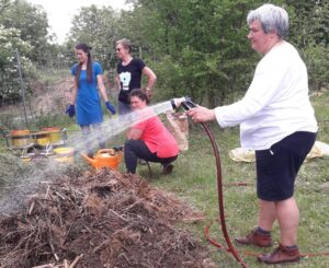 eine Person spritzt mit einem Wasserschlauch auf einen Komposthaufen, dahinter drei weitere Frauen mit Gartenhandschuhen