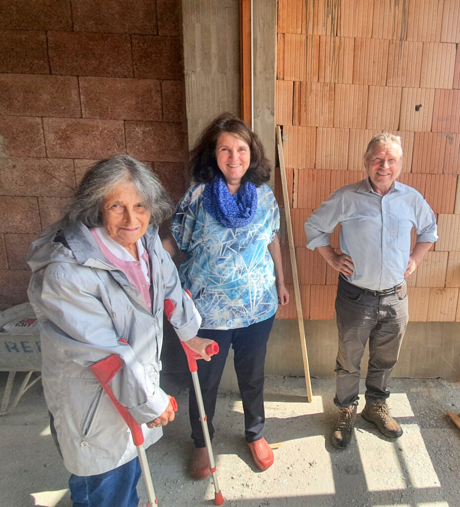 eine betagte Dame mit zwei Krücken, eine ältere Frau und ein älterer Mann stehen vor einer Ziegelwand auf der Baustelle.