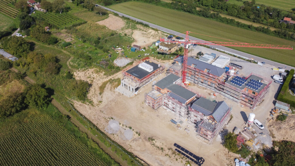 Drohnenbild von der Baustell: drei im Bau befindliche Gebäude, auf einem eine PV-Anlage, im Vordergrund ein Tipi, rundherum Baustelle und darum Grünflächen
