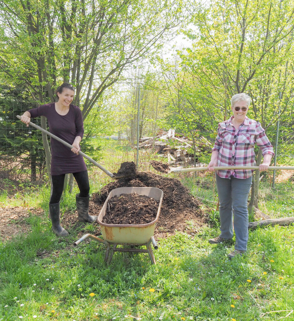 eine junge Frau und eine ältere stehen links und rechts von einem Komposthaufen und schaufeln in die Scheibtruhe, die zwischen ihnen steht. Sie blicken lachend in die Kamera. Im Hintergrund Bäume und Büsche