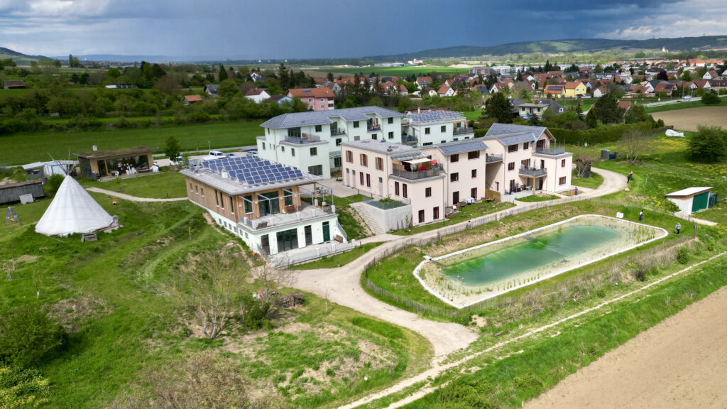 die Gebäude aus der Vogelperspektive: ein zweistöckiges Haus teilweise mit Holzverkleidung und PV-Anlage, ein Teich, dahinter ein rötliches Wohngebäude mit drei Stockwerken, und dahinter ein grünliches Wohngebäude mit drei Stockwerken. Alles in Hanglage und im Grünen.