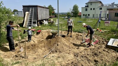 3 Erwachsene und ein Kleinkind stehen in einer großen Sandgrube und graben um. Das Kind hält einen Kübel hoch. Im Hintergrund ein Bauwagen auf einer Wiese und ein Wohnhaus.