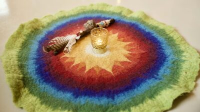 ein runder Filzteppich mit unterschiedlichen Farben und einer Sonne in der Mitte. Im Mittelpunkt der Sonne steht ein Glas mit einem brennende Teelicht. Daneben liegt ein hölzerner Redestab, mit Federn geschmückt.