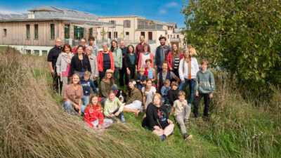 Eine Gruppe von Menschen verschiedener Altersstufen, zwei Hunde und ein Huhn stehen auf einer Wiese auf einer Anhöhe. Dahinter sind moderne Gebäude zu sehen, ein Baum im Anschnitt.