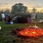 die Glut eines Feuers, im Hintergrund legen Menschen Decken auf ein Holzgerüst der Schwitzhütte