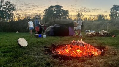 die Glut eines Feuers, im Hintergrund legen Menschen Decken auf ein Holzgerüst der Schwitzhütte