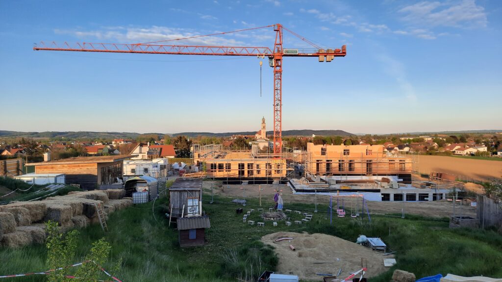 eine Baustelle mit einem halbfertigen Ziegelrohbau, darüber ein Kran vor blauem Himmel